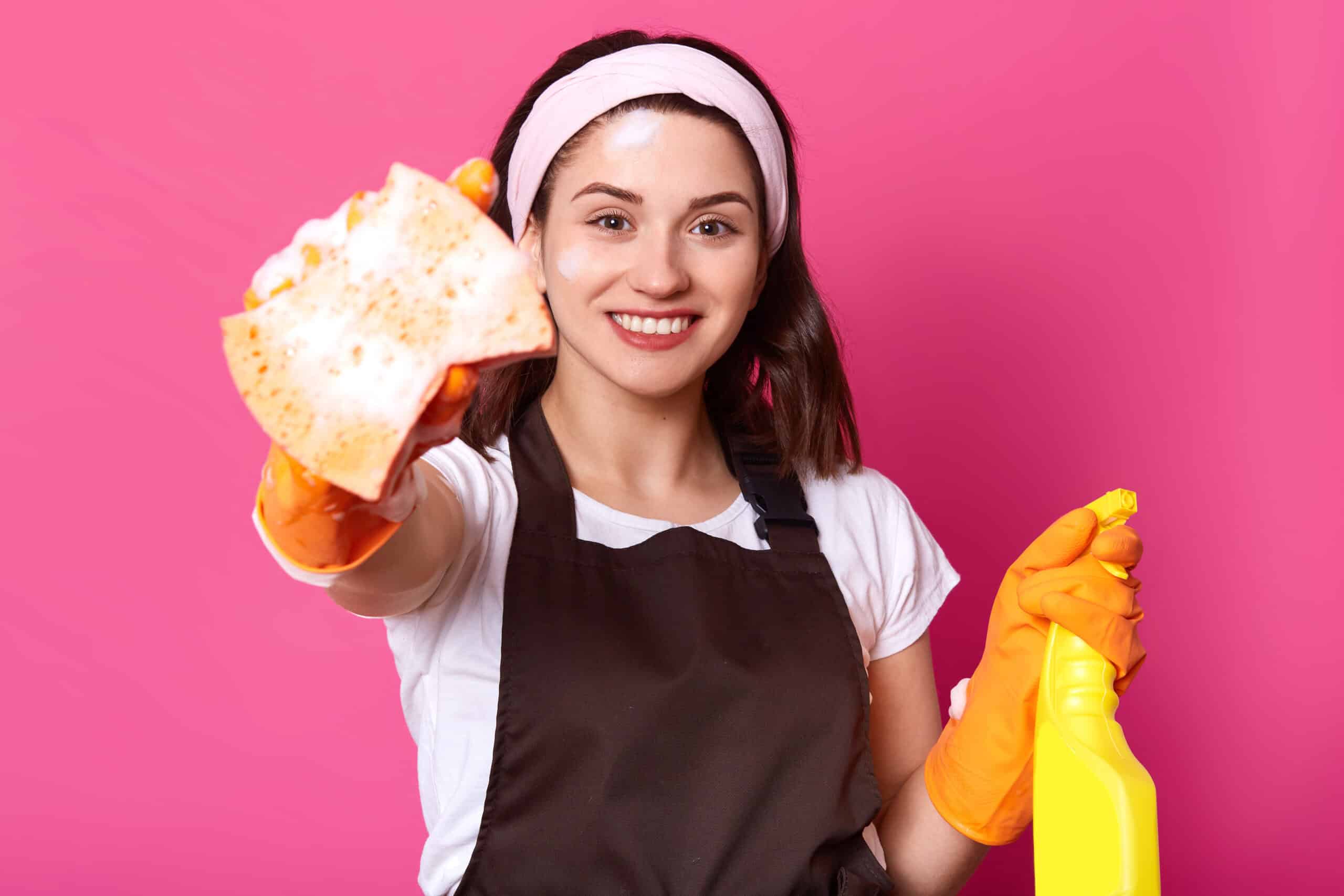 Indoor shot of happy Caucasian young woman in white casual t shirt, hairband, brown apron, holds sponge and cleaning detergent, ready to do Spring Cleaning